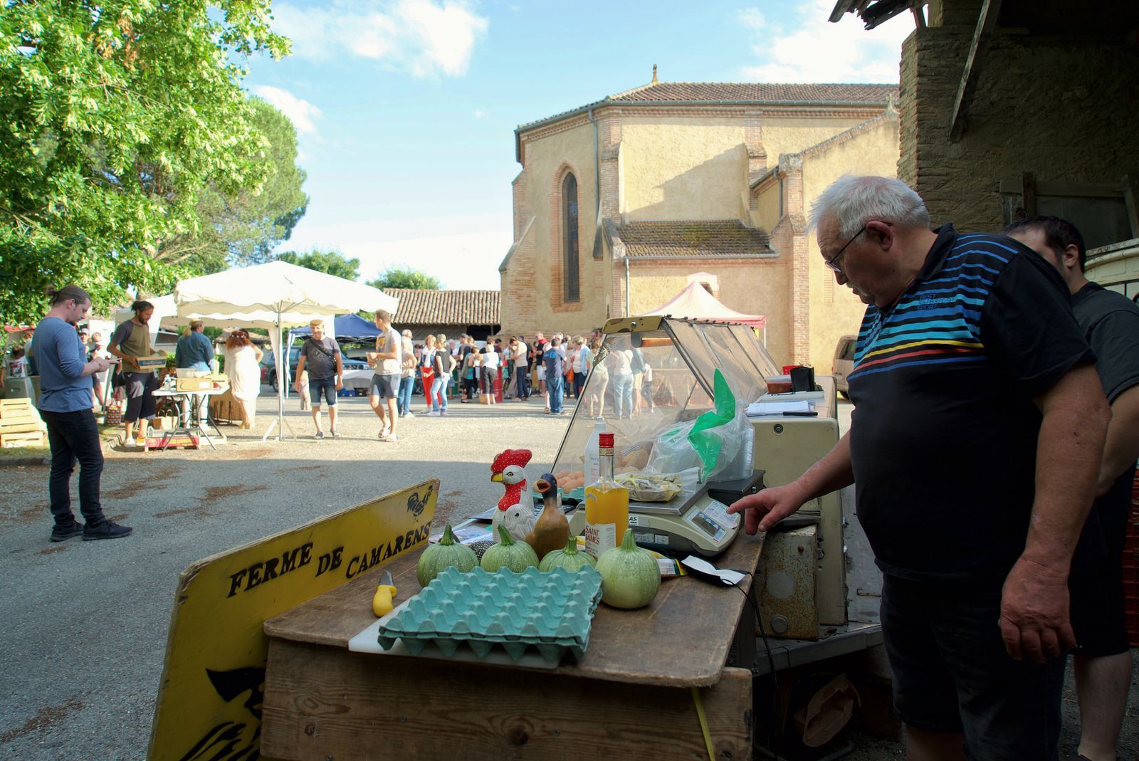 Marché animé sur la place de Noilhan : un étal de fruits et d’œufs au premier plan, l’église en arrière-plan